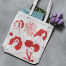 Tan canvas tote adorned with four women in red vinyl and floral detail, carrying a magazine, photographed on grey background.