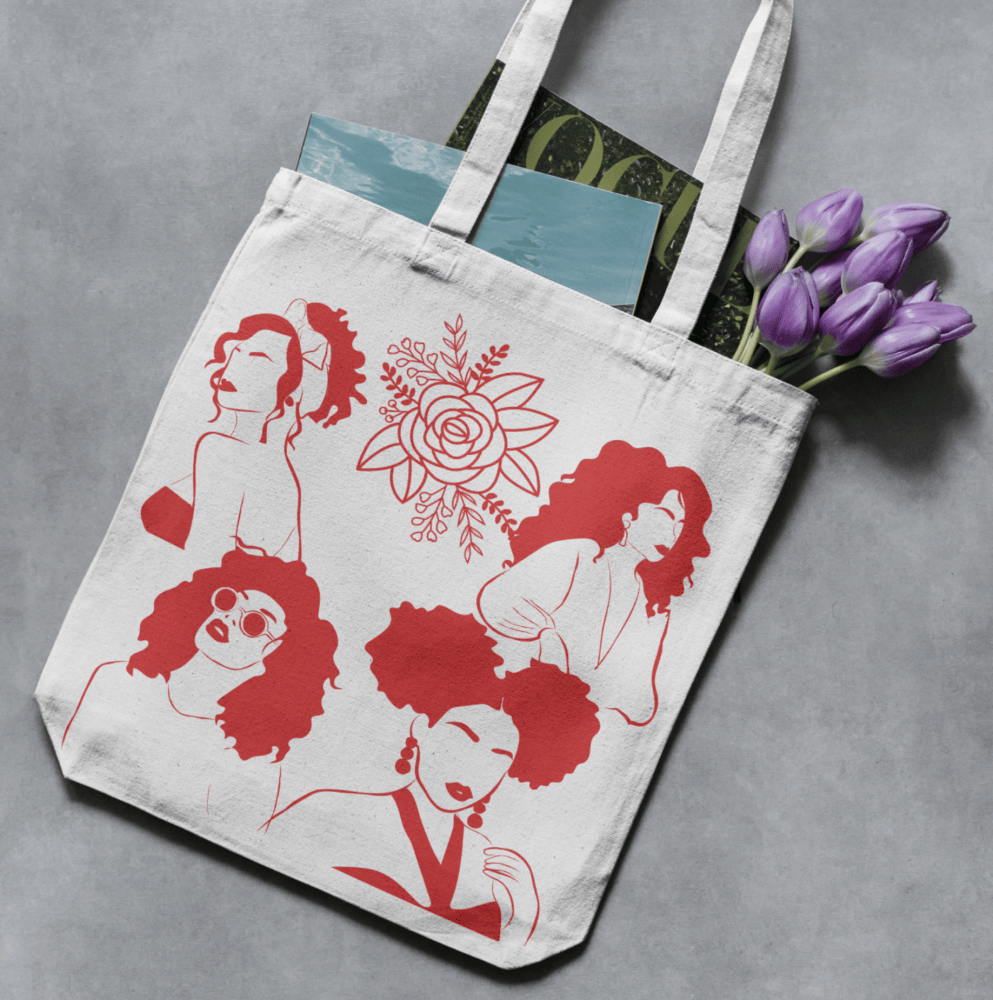 Tan canvas tote adorned with four women in red vinyl and floral detail, carrying a magazine, photographed on grey background.
